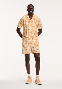 Man staat met zijn handen achter zijn rug, gekleed in een lichtbeige shirt met korte mouwen en bijpassende korte broek met een oranje bloemenprint, en oranje hoge sneakers.