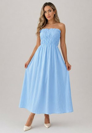 Femme aux longs cheveux blonds porte une robe longue bleu clair sans bretelles et des talons blancs, debout devant un fond clair uni.