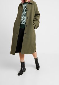 Olivgrüner Wollmantel mit Knöpfen, großem Kragen und seitlichen Taschen, übereinander getragen über einem gestreiften Strickpullover und einem schwarzen Rock, kombiniert mit schwarzen Stiefeletten.