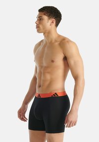 Černé boxerky s oranžovým pasem, na kterém je černé logo se třemi pruhy. Hladká látka, přiléhavý střih a délka do půlky stehna.