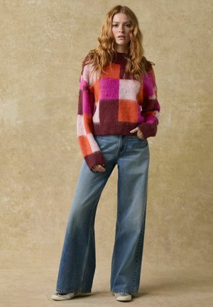 Kolorowy sweter w wzory w odcieniach różowego, pomarańczowego i brązowego; zestawiony z jasnoniebieskimi dżinsami w stylu dzwonów oraz beżowymi butami. Teksturowany materiał i luźny krój.