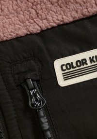 Zoom na zadrgo črne jakne in bež nalepko z besedilom "COLOR K" na črnem blagu pod teksturiranim rjavim flisom.