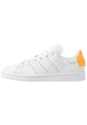 Zapatilla blanca de cuero de corte bajo con cordones blancos, rayas laterales perforadas y pestaña naranja en el talón, con un logo grabado en la suela.