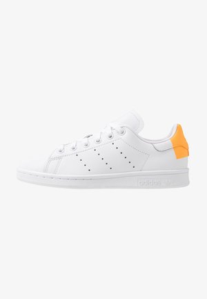 Zapatilla blanca de cuero de corte bajo con cordones blancos, rayas laterales perforadas y pestaña naranja en el talón, con un logo grabado en la suela.