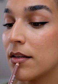 Un lápiz de labios de un tono marrón claro se sostiene cerca de los labios de una persona. Piel suave con un brillo sutil; cejas definidas y delineador de ojos.