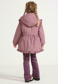 Roze gewatteerde jas met ruches aan de randen, verstelbare taille en lange mouwen. Gepaarde met paarse corduroy broek en zwarte schoenen.