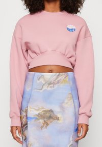 Persona che indossa un cropped sweatshirt rosa con la scritta "Good Times" e una gonna alta con una stampa di cherubini rinascimentali e nuvole.