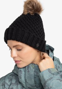 Gorro de punto negro con un diseño texturizado y un pompón de piel sintética en la parte superior. Cuenta con un puño acanalado y un pequeño detalle de logotipo en el lado.
