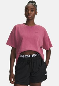Vrouw met gevlochten haar draagt een losse roze cropped T-shirt en zwarte Under Armour mesh sportshorts met haar handen in de zakken.