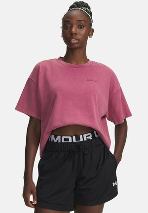 Kvinna med flätat hår bär en lös rosa kortärmad tröja och svarta Under Armour mesh sportshorts med händerna i fickorna.