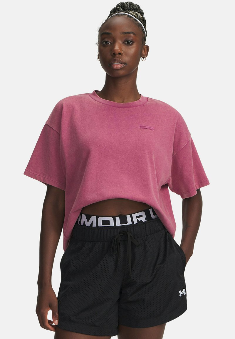 Vrouw met gevlochten haar draagt een losse roze cropped T-shirt en zwarte Under Armour mesh sportshorts met haar handen in de zakken.