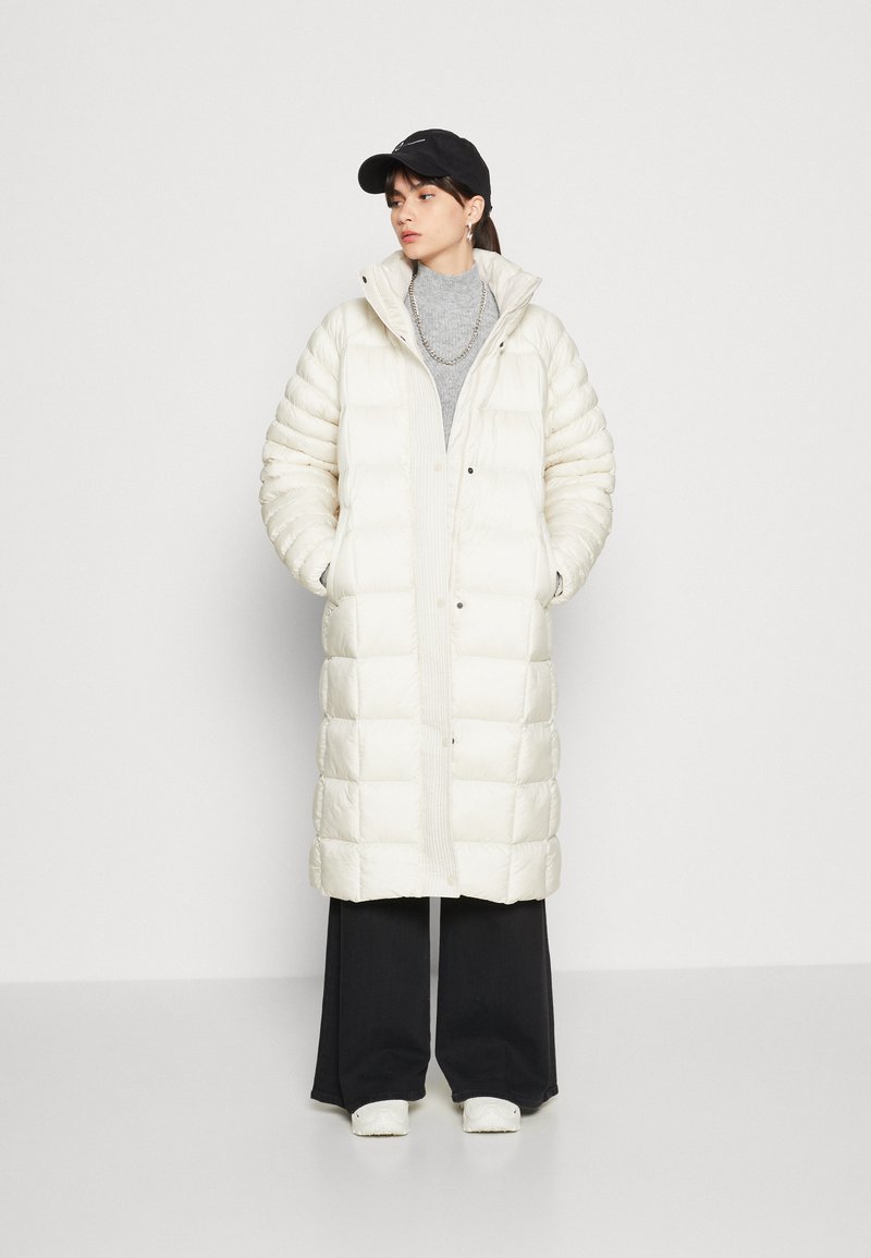 Manteau long rembourré blanc avec un col haut, des boutons-pression, des détails à côtes verticales, associé à un pantalon large noir et une casquette noire.