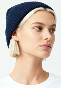 Cappellino a coste blu navy, con trama lavorata a maglia, design aderente, indossato su capelli biondi corti, maglietta bianca visibile sotto.