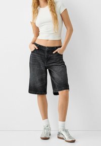 Black denim bermuda shorts with a relaxed fit, 5-pocket design, and cuffed hems, paired with a fitted white t-shirt and silver sneakers.