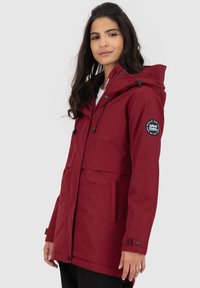 Vrouw in een bordeauxrode hoodie met voorzakken en een rond logo-embleem op de linkermouw, tegen een effen achtergrond.