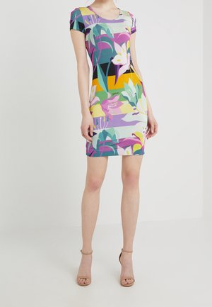 Femme portant une robe à manches courtes, longueur genou, avec un motif floral et abstrait coloré, associée à des talons ouverts beige.