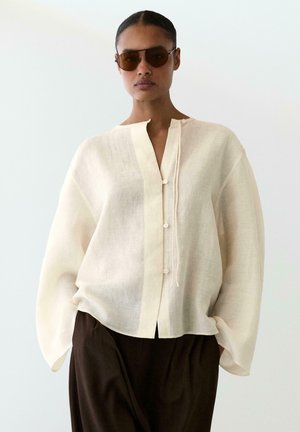 Žena nosící nadměrně velkou průsvitnou bílou halenku se zapínáním na knoflíky, volné rukávy, tmavě hnědé kalhoty a hnědé sluneční brýle, stojící na bílém pozadí.