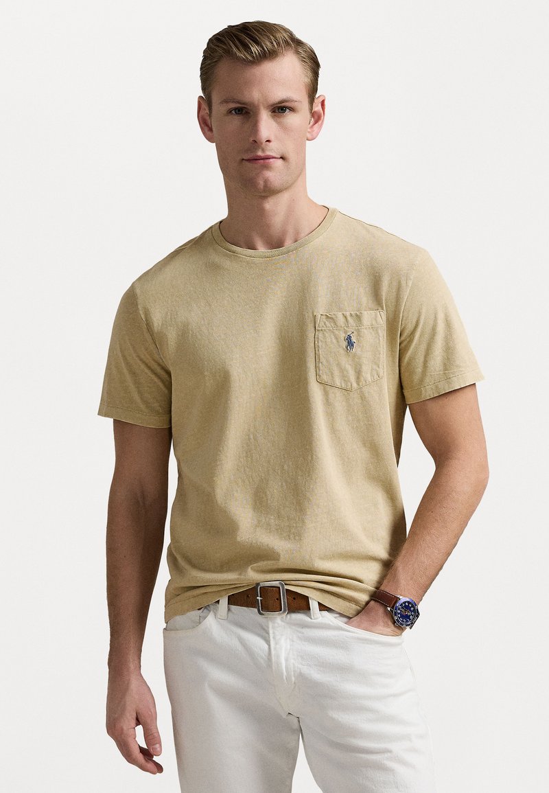 Μπεζ βαμβακερό μπλουζάκι με κοντό μανίκι, στρογγυλή λαιμόκοψη, μπροστινή τσέπη και μικρό λογότυπο στην τσέπη. Συνδυασμένο με λευκά παντελόνια και ρολόι.