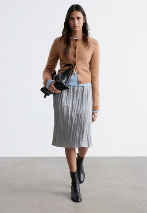 Kvinde går iført en tanfarvet cardigan med knapper, blå sweater, grå midi-nederdel, sorte ankelstøvler og holder en sort tekstureret håndtaske.