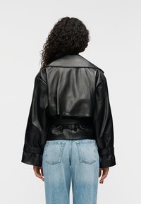 Μαύρο δερμάτινο μπουφάν με κοντή γραμμή, φαρδιά γιακά και ζώνη στη μέση· διαθέτει φαρδιά μανίκια και λεία υφή. Φορεμένο πάνω από ανοιχτόχρωμο μπλε τζιν.