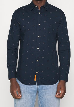 Mand iført en navyblå skjorte med små diamantmønstre og lyseblå jeans, stående mod en ensfarvet baggrund.