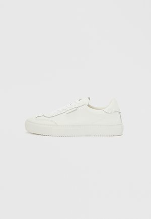 Hvid lav sneaker med bølgede kanter, snørelukning og flad gummisål på en ensfarvet hvid baggrund.