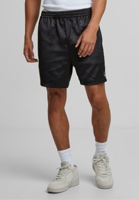 Sorte atletiske shorts med elastisk talje, prydet med et subtilt mønster og et lille logo nederst. Bæres med hvide sokker og sneakers.