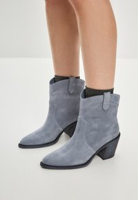 Botas de tobillo de ante gris con punta afilada, tacón apilado y diseño con puño, que presentan una textura sutil y mínimos detalles de costura.