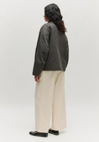 Oversized donkergrijze jas met een rechte snit, gecombineerd met wijde beige broeken en zwarte platte schoenen, gezien vanaf de achterkant.