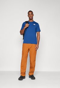 Μπλε κοντομάνικο μπλουζάκι με λογότυπο στο στήθος, συνδυασμένο με πορτοκαλί τσιγαράκι παντελόνι και μαύρα παπούτσια. Το μοντέλο κρατάει μια μαύρη τσάντα πλάτης.