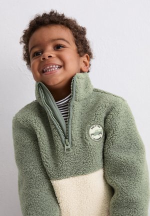 Lächelndes, junges Kind trägt eine grüne und crèmefarbene Fleecejacke mit einem halben Reißverschluss und einem runden Logo-Patch auf der Brust vor einem schlichten Hintergrund.