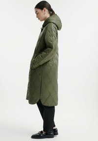 Cappotto lungo trapuntato verde oliva con cappuccio, caratterizzato da tasche laterali, una texture liscia e un orlo curvato, indossato sopra jeans neri e scarpe.