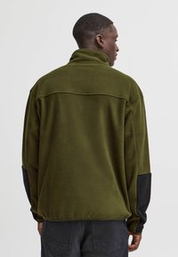 Sudadera de forro polar verde oliva con un alto cuello y detalles negros en las mangas. Textura suave, corte holgado, detalle de costura mínima.