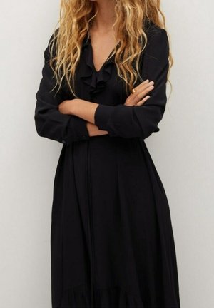 Femme aux longs cheveux blonds bouclés portant une robe noire à manches longues avec des détails à volants, les bras croisés devant un fond uni.