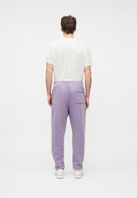 Άνδρας που στέκεται με την πλάτη γυρισμένη, φοράει λευκό κοντομάνικο πουκάμισο, λιλά φόρμα με πίσω τσέπη και λευκά αθλητικά παπούτσια σε λευκό φόντο.