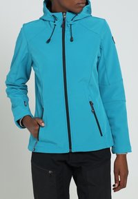 Personne portant une veste à capuche turquoise avec fermeture éclair, cordons et poches zippées, assortie à un pantalon noir.