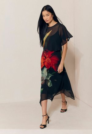 Vrouw met lang zwart haar, gekleed in een zwarte jurk met doorschijnende wapperende mouwen en een grote rood-witte bloemenprint, lopend op zwarte hoge hakken sandalen.