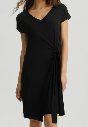 Femme portant une robe noire à manches courtes avec un col en V et un détail de nœud froncé à la taille, debout devant un fond uni.