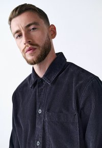 Dunkelblaues Cordhemd mit strukturiertem Kragen, durchgehender Knopfleiste und einer einzelnen Brusttasche, mit vertikal geribter Textur.