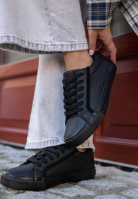 Czarne skórzane sneakersy z teksturowaną boczną panelem, płaskimi sznurowadłami i gumowymi podeszwami. Noszone z szarymi dżinsami, co podkreśla swobodny styl.