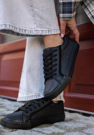 Czarne skórzane sneakersy z teksturowaną boczną panelem, płaskimi sznurowadłami i gumowymi podeszwami. Noszone z szarymi dżinsami, co podkreśla swobodny styl.