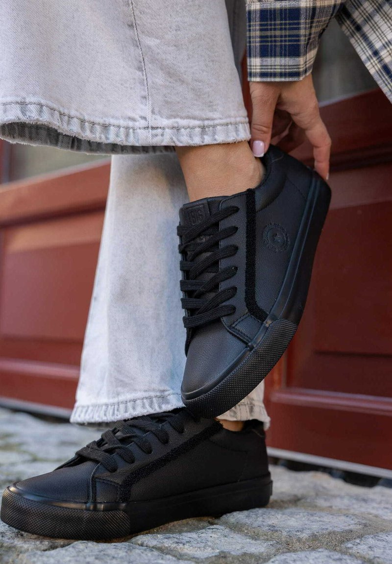 Czarne skórzane sneakersy z teksturowaną boczną panelem, płaskimi sznurowadłami i gumowymi podeszwami. Noszone z szarymi dżinsami, co podkreśla swobodny styl.