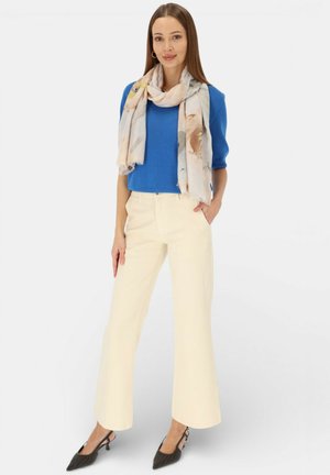 Kobieta stojąca, ubrana w niebieski sweter z krótkim rękawem, jasno kolorowy szal, kremowe szerokie spodnie i czarne sandały na obcasie z odkrytą piętą, z ręką w kieszeni.