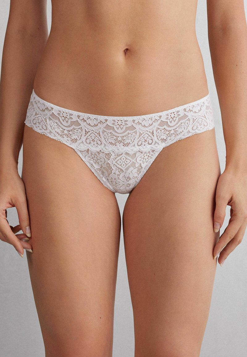String en dentelle blanche avec un motif floral, des bords festonnés et une taille lisse. Semi-transparent avec des détails délicats.