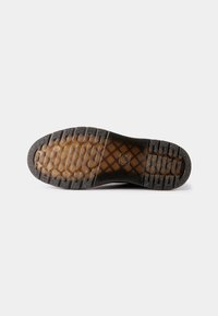 Semelle d'une botte noire avec un motif de bande de roulement marron transparent et des éléments de grip texturés sur fond blanc.