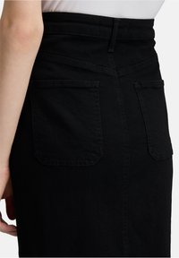 Femme portant une jupe en denim noir taille haute avec deux poches arrière, associée à un haut blanc rentré, la main reposant à proximité.