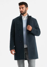 Cappotto di lana blu scuro con sei bottoni, colletto alto e una texture liscia, indossato sopra un maglione azzurro chiaro e pantaloni grigi.