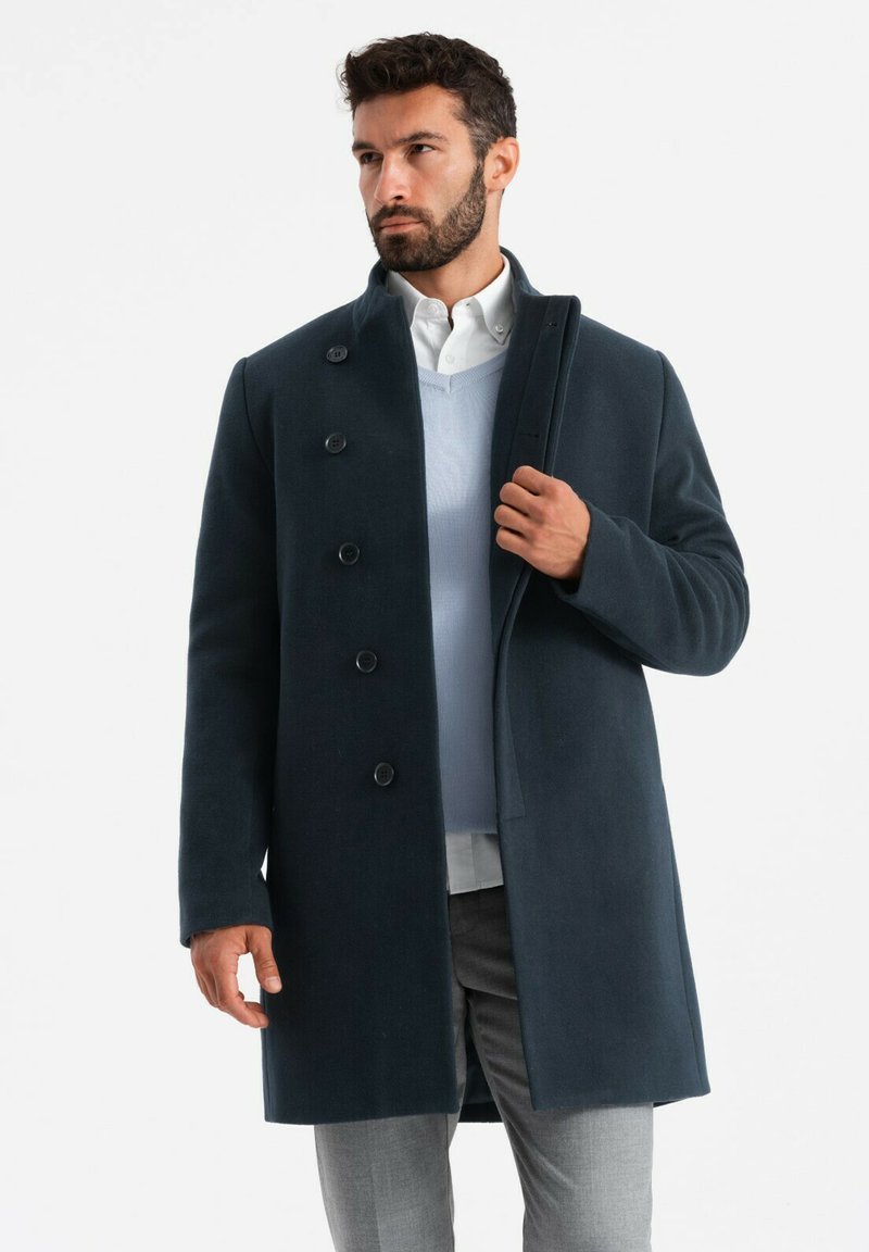 Cappotto di lana blu scuro con sei bottoni, colletto alto e una texture liscia, indossato sopra un maglione azzurro chiaro e pantaloni grigi.