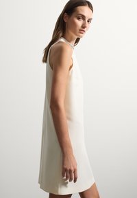 Vestido sin mangas de color crema con una silueta recta, que presenta un escote texturizado y una tela suave. Tiene un dobladillo plano y detalles de abertura lateral.