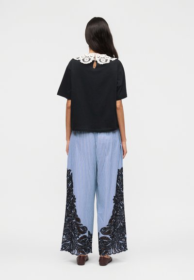 Γυναίκα που φοράει μαύρο τοπ με κοντά μανίκια και λευκό δαντελένιο γιακά, καθώς και ανοιχτό μπλε παντελόνι με φαρδιά πόδια και μαύρη δαντελένια κεντητική, στέκεται με την πλάτη προς τα εμπρός.